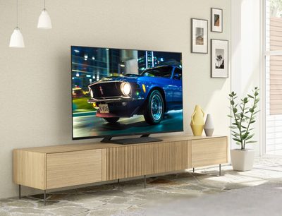 Panasonic TX-65HX800E-fladskærm, der viser en blå Ford Mustang fra 1968 på en nattebelyst gade, stående på et lyst trækabinet