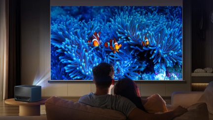 4K ultraīsā fokusa lāzera projektors, klaunu zivju attēls uz ekrāna, divu cilvēku aizmugures skats, sēžot uz dīvāna