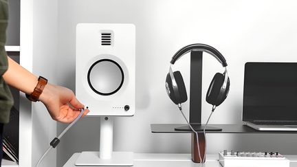 Vedere frontală a unei incinte rectangulare albe mate din plastic, așezată pe un suport, traversată de un tweeter cu lamele și un woofer mare cu ramă neagră, o mână conectând un cablu audio împletit gri, cu o pereche de căști negre și argintii agățate de un suport metalic în dreapta și un laptop parțial vizibil.