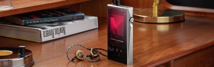 Lettore audio portatile color argento con schermo acceso, cuffie nere e dorate, libri, lampada dorata, scrivania in legno