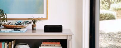 Čierny stereo hi-fi zosilňovač Sonos Amp položený na bielej poličke vedľa kníh a zelenej izbovej rastliny v kvetináči