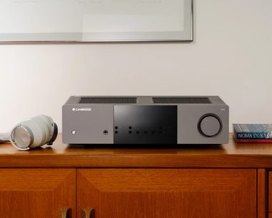 Cambridge Audio EXA100 integrált sztereó hi-fi erősítő, előlről nézve, szürke téglalap alakú ház, jobboldalt kör alakú gomb, tetején szellőzőrács, baloldalt fehér fejhallgató, jobboldalt könyvek halmaza