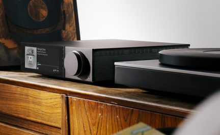 Amplificatore audio nero in metallo con pannello frontale dotato di schermo a colori e ampia manopola del volume, lato scanalato per la ventilazione, vista in tre quarti dall’alto leggermente inclinata