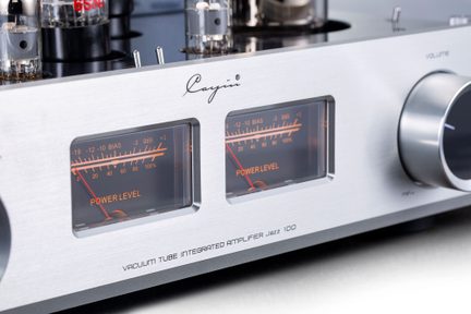 Stříbrný hi-fi lampový zesilovač z kartáčovaného hliníku, pohled zepředu z tří čtvrtin, přední panel s dvěma retro osvětlenými VU metry v oranžové barvě, viditelné lampy na vrchní části a velký chromovaný ovládací knoflík hlasitosti vpravo