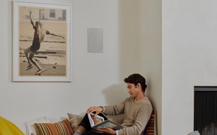 Vit vägg med vit träram och väggmonterat högtalargaller i metall, man som läser på soffa med randiga kuddar