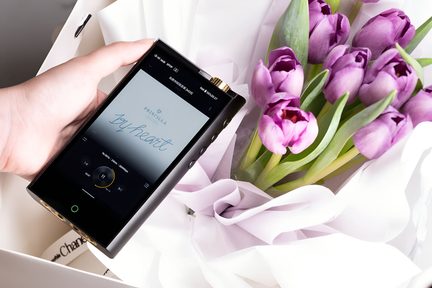 Gros plan oblique sur un lecteur audio portable noir en métal avec molette dorée et boutons latéraux, tenu dans une main gauche, à côté d’un bouquet de tulipes violettes enveloppées de papier blanc