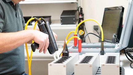 Zbliżenie trzech prostokątnych, metalicznych, szarych modułów testowych z owalnymi otworami, wraz z otwartą, wyposażoną, szarą walizką, żółtymi przewodami w osłonie z sprężynami oraz czarnymi plastikowymi uchwytami sond, wykonane z perspektywy trzech czwartych.