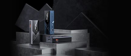 Drie rechthoekige elektronische behuizingen van matzwart en nachtblauw metaal, voorzien van een digitaal frontdisplay, een gegraveerd X-motief en een rode USB-C-poort, driekwart in beeld gebracht op donkere treden
