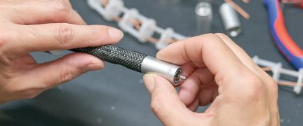 Hænder holder et sort kabel med sølvfarvet cylindrisk metalstik, sløret baggrund med værktøj og komponenter