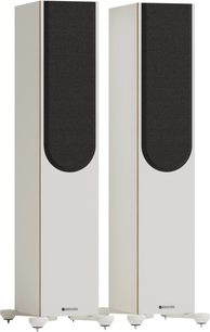 İki adet Monitor Audio Bronze 300 7G kolon hoparlör, mat beyaz ahşap kasada, siyah ön ızgara, kenarlarda bronz şerit ve sivri ayaklarla, üç çeyrek ön görünüm