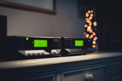 Cyrus 82 DAC QXR sort hi-fi stereoforstærker, nærbillede forfra, grønne displays tændt, sølvfarvede knapper, slørede lys i baggrunden