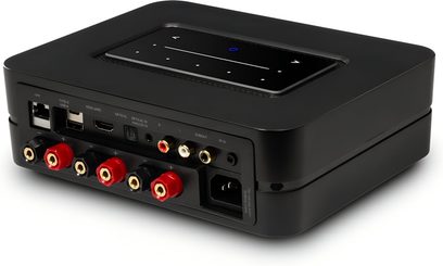 Інтегрований стерео підсилювач Hi-Fi BlueSound Powernode 2025 (N331) чорного кольору, корпус з матової пластмаси з глянцевою сенсорною панеллю зверху, ряд мережевих портів USB HDMI оптичний RCA та червоні/чорні клемні колодки для динаміків на видноті, задній вид у перспективі