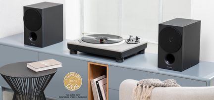 Silberne und schwarze Vinyl-Plattenspieler-Platine aus Metall und Kunststoff mit Schallplatte auf dem Plattenteller, leicht von oben frontale Ansicht, eingerahmt von zwei rechteckigen schwarzen Lautsprechern mit matter Gehäusebeschichtung und kreisförmigem Gitter