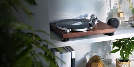 giradischi in legno visto di tre quarti posizionato su uno scaffale bianco con disco nero, altoparlante sferico, libri, piante
