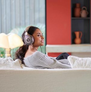 Ung kvinde siddende på en hvid sofa, iført en grå sweater, lyttende til musik med en sølvfarvet hovedtelefon
