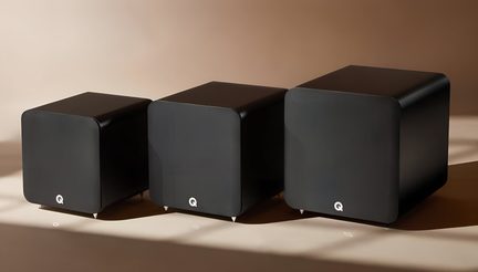 Three matte black cubic enclosures, front panel in black acoustic fabric with a discreet white symbol, rounded edges, metallic conical feet, arranged from smallest to largest in a three-quarter front view