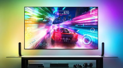 Pantalla plana rectangular mostrando un juego de carreras colorido, 2 barras luminosas verticales negras, mueble con objetos debajo.