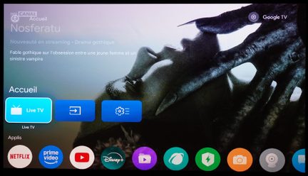 Svart platt skärm med plastkant och glasytta, sedd framifrån, som visar Google TV-gränssnittet med en stor mörk filmillustration och en rad med färggranna appikoner längst ner