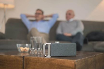 Szürke hordozható Bluetooth-hangszóró ezüst fogantyúval, dohányzóasztalon elhelyezve