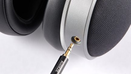 Nahaufnahme eines Kopfhörers in Metallic-Grau, Ohrpolster aus schwarzem Kunstleder, schwarze Stoffabdeckung und vergoldeter 3,5-mm-Klinkenanschluss mit ausgerichtetem schwarzem Audiokabel