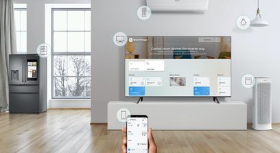Televizor Samsung QE65Q9F 2018 v moderním obývacím pokoji s okny, šedou lednicí, dřevěnou podlahou a rukou držící telefon s aplikací SmartThings