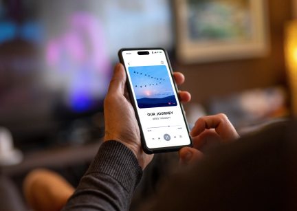 Svart smarttelefon holdt i hånden, skjermen viser lydspilleren for sporet Our Journey med illustrasjon av blå himmel og fugler