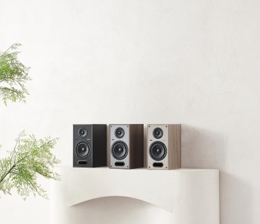 Tři regálové reproduktory v černém dřevě, ořechu a světlém dubu, šedá čelní stěna, výškový reproduktor, basový reproduktor a obdélníkový bassreflexový otvor