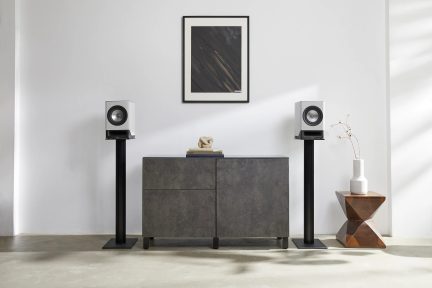 Vista frontal, 2 altavoces blancos sobre soportes negros, mueble gris, cuadro abstracto, jarrón blanco sobre mesa de madera