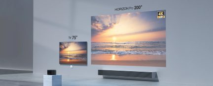 Minimalistyczna, szara przestrzeń z przodu, z dużym, 200-calowym ekranem projekcyjnym 4K wyświetlającym zachód słońca, mniejszym, 75-calowym telewizorem po lewej stronie, matowo-czarnym, metalowym projektorem w kształcie sześcianu ustawionym na białym marmurowym postumencie oraz ciemnoszarą, poziomą belką na podłodze.