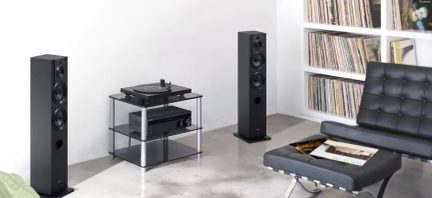 Två svarta högtalare, glasmöbel med skivspelare och förstärkare, hylla med vinylskivor, svart stoppad fåtölj