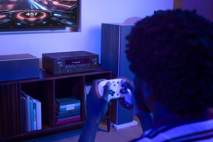 Sort Denon hjemmebiografiforstærker med frontdisplay, mellem to højttalere under et gaming-tv