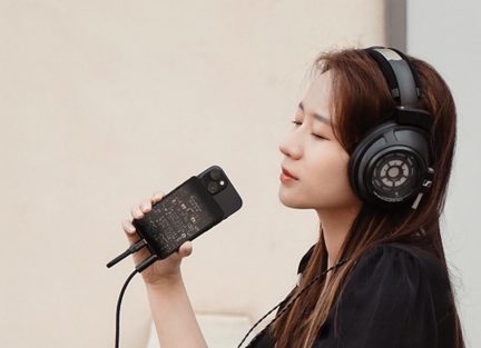 Căști audio Sennheiser circumaurale negre conectate la smartphone cu amplificator DAC portabil, femeie ascultând muzică