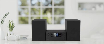 Kompaktní černá hi-fi sestava Kenwood se dvěma reproduktory a centrálním displejem, pohled zepředu na bílém stole