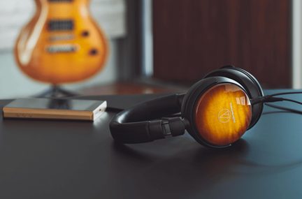 Sort over-ear hovedtelefoner i sort med polstrede ørepuder og træskaller i orange farveovergang med hvidt logo, vist i trekvarts vinkel placeret på en mørk overflade