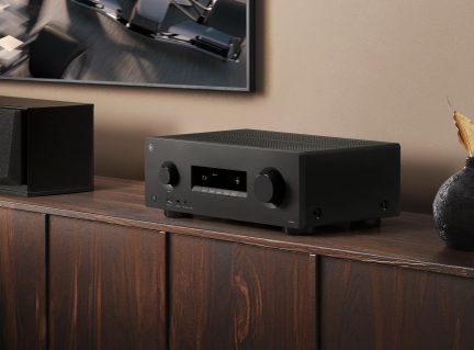 Svart Yamaha nettverksreceiver med sentralt display og doble dreieknapper, plassert på et salongmøbel i tre