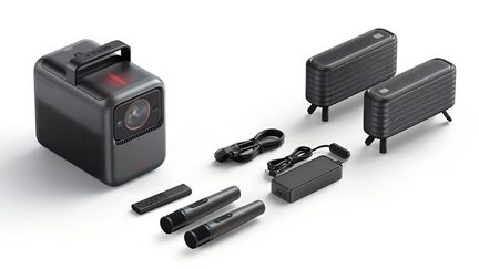 Matte black portable projector in plastic with a top handle and a red-ringed lens, three-quarter front left view, accompanied by two black cylindrical microphones, two black mesh speakers on short stands, a flat remote control, and a power supply unit with black cables