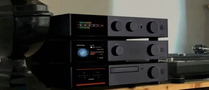Trei dispozitive audio negre stivuite, cu butoane circulare, ecrane luminoase și un player de discuri, pe o suprafață de lemn