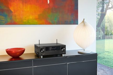 Sort audio-/videomodtager placeret på et sort møbel, rød skål og hvid lampe ved siden af, farverigt abstrakt maleri på væggen bagved