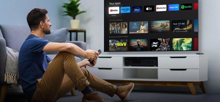 Soggiorno contemporaneo luminoso, grande televisore a schermo piatto nero visto di fronte che mostra un’interfaccia di streaming, posizionato su un mobile basso in legno bianco con gambe inclinate color rovere, uomo bruno con t-shirt blu e pantaloni beige seduto a terra che tiene un telecomando