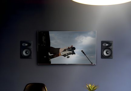 Muro grigio, TV a schermo piatto in vetro tra due casse murali nere, visualizzazione di un manico di chitarra e di un microfono