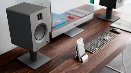 Birou din lemn maro cu vene, vedere din trei sferturi ușor de sus, cu monitor din aluminiu gri deschis, boxă cubică gri închis mat pe suport în prim-plan, a doua boxă asortată în spate, tastatură neagră, mouse gri și smartphone pe suport metalic