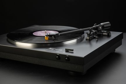 Tourne-disque noir et gris en métal et plastique, plateau argenté portant un disque vinyle noir, bras de lecture noir avec cellule jaune étendu vers le centre, deux boutons avant start et stop visibles, vue de trois-quarts avant