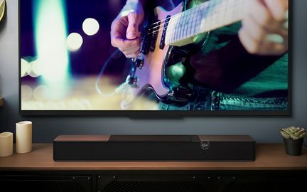 Schwarze Klipsch-Soundbar mit Kupferelementen auf TV-Möbel, unter großem Bildschirm mit E-Gitarristen