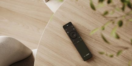 Telecomandă neagră We by Loewe așezată pe o măsuță rotundă cu aspect de lemn deschis, văzută de sus