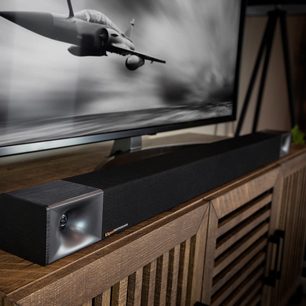 Bară de sunet Klipsch Cinema 600 neagră pe mobilier din lemn, vedere frontală de aproape, mână ținând o telecomandă îndreptată spre bara de sunet