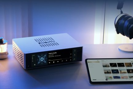Kompakter Netzwerk-Audioplayer aus silberfarben eloxiertem Aluminium, Dreiviertelansicht von vorn, komplett verglaste Front mit großem schwarzem Farbdisplay und Gehäuse mit Reihen kreisförmiger Löcher, die auf der Oberseite und der linken Seite ein M-Muster bilden