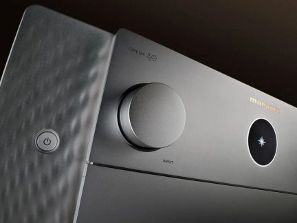 Primer plano en tres cuartos de un amplificador gris de metal cepillado, con un gran botón de entrada, pantalla redonda luminosa y panel lateral texturizado