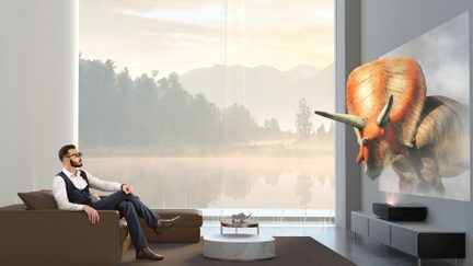 Mand siddende på en brun sofa, der ser en holografisk projektion af en triceratops i en moderne stue med store vinduer