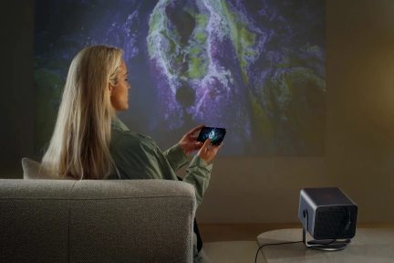 Videoprojetor cinzento num suporte, colocado sobre uma mesa, com projeção na parede e mulher a segurar um smartphone