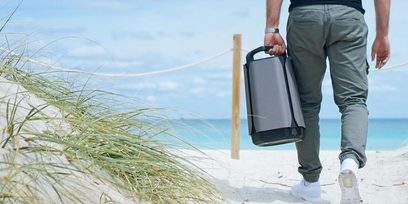 Mand, der går på en strand, iført grå bukser og hvide sneakers, bærende en sort kuffert i hånden